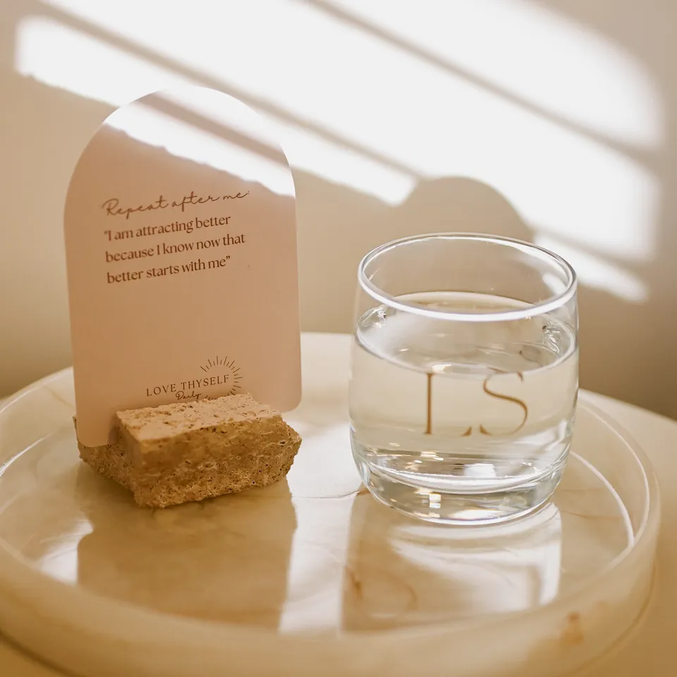 A glass of water labeled "LS" sits on a round wooden surface next to a card with an inspirational quote.