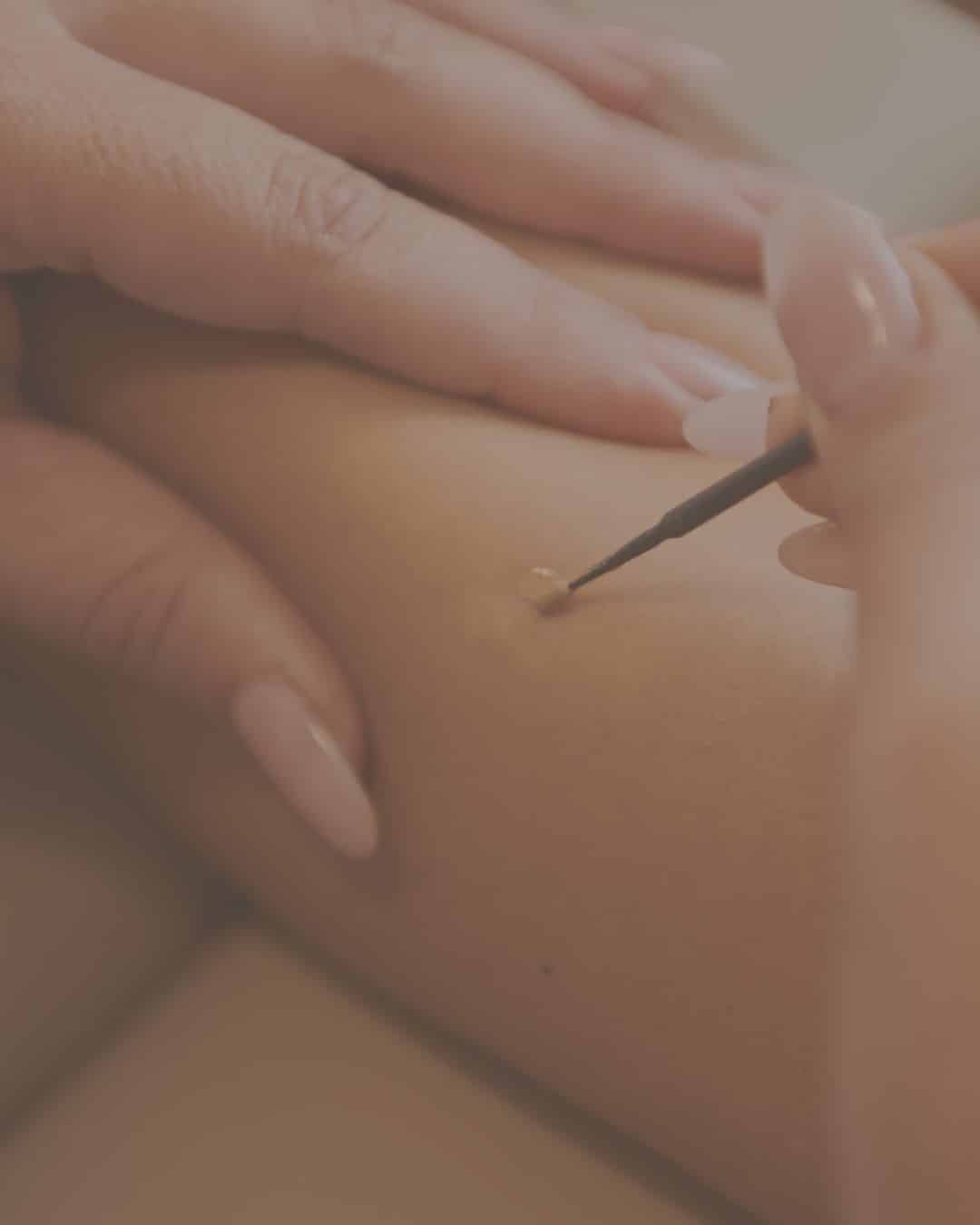 A person uses a small brush to apply a drop of beige liquid makeup to another person's skin.