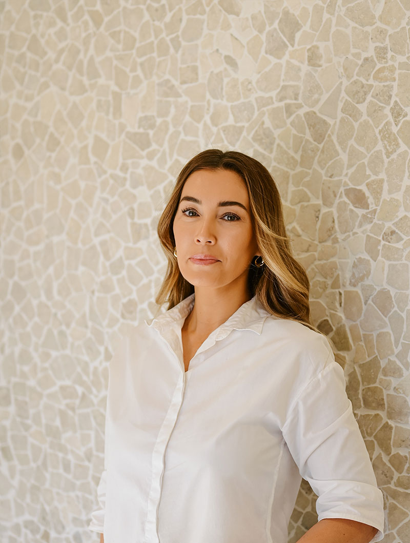 A woman with long brown hair wearing a white button-up shirt stands against a light-colored mosaic-patterned wall, looking at the camera.