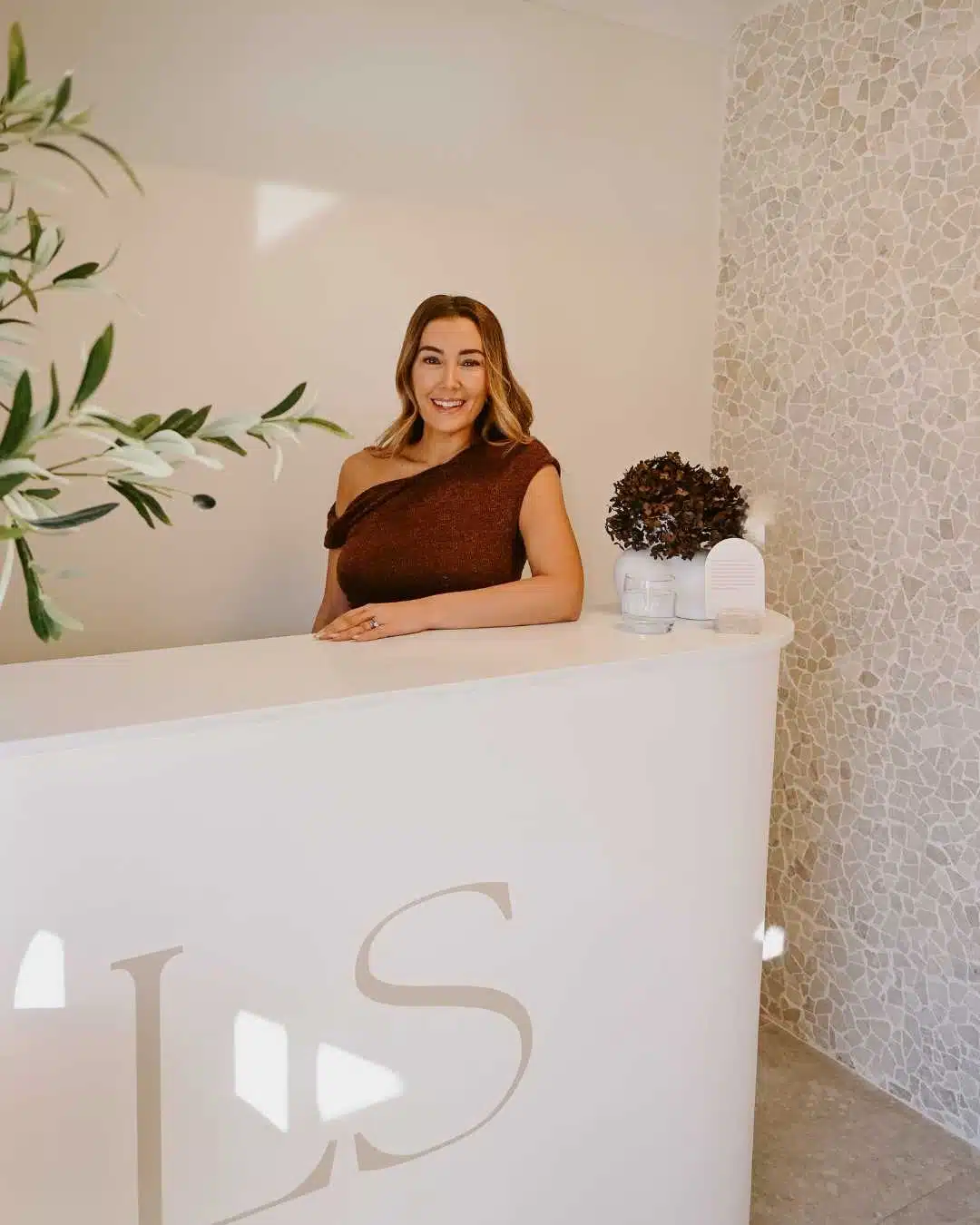 A woman stands behind a white reception desk with "LS" on the front, in a bright room with mosaic tile walls and decorative plants. I Stretch Mark & Scar Treatment Perth Clinic