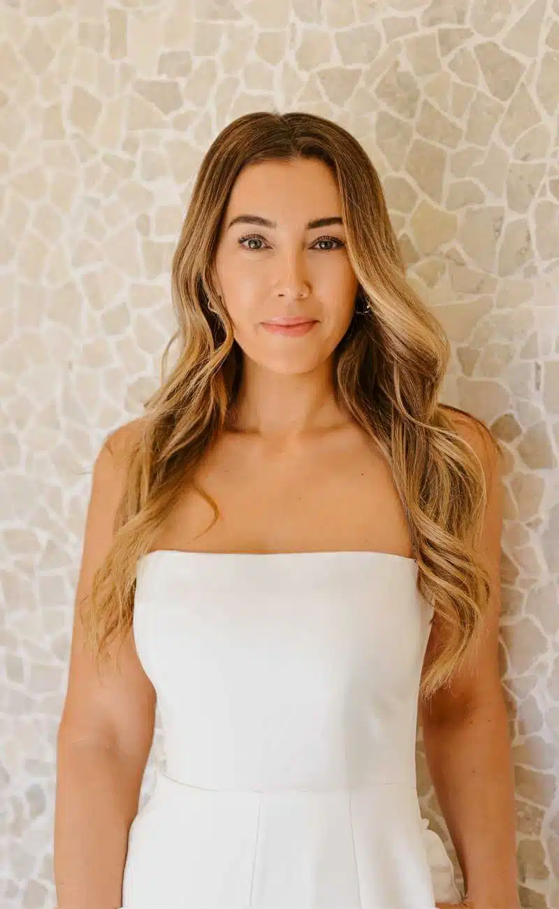 Woman with long, wavy hair wearing a white strapless dress stands in front of a light mosaic tile wall, looking at the camera. I Stretch Mark & Scar Treatment Perth Clinic