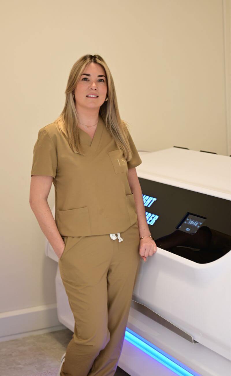 A woman in tan medical scrubs stands next to a white medical or spa machine in a minimalist room.