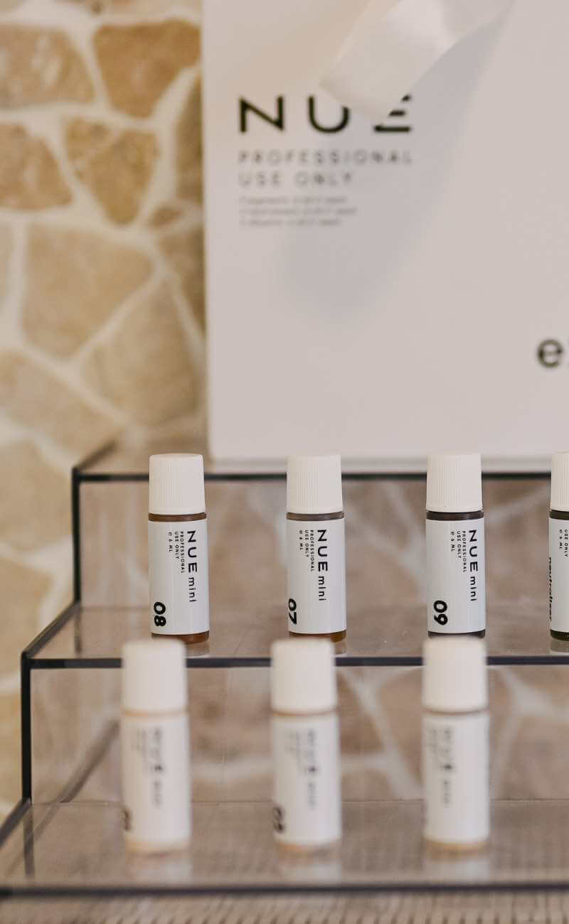 Small white bottles labeled "NUE" are arranged on a tiered clear display stand, with a white sign in the background that reads "NUE Professional Use Only. I Stretch Mark & Scar Treatment Perth Clinic