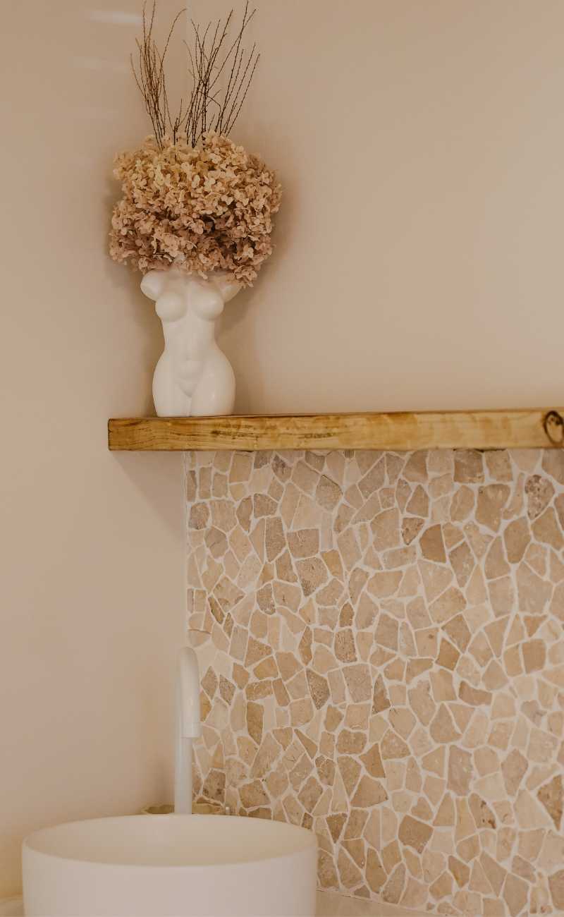 A white vase shaped like a female torso with dried flowers sits on a wooden shelf above a tiled mosaic wall and a round white sink. I Stretch Mark & Scar Treatment Perth Clinic