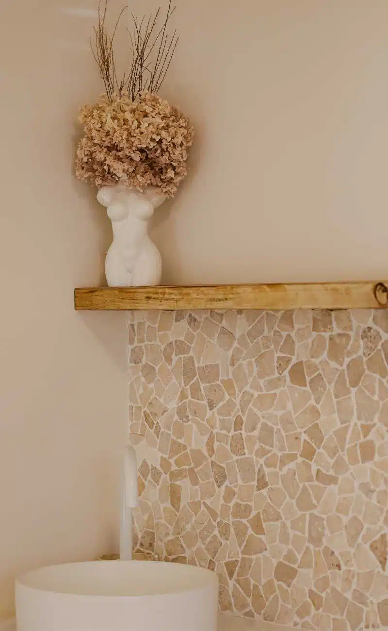 A white vase shaped like a female torso with dried flowers sits on a wooden shelf above a tiled mosaic wall and a round white sink. I Stretch Mark & Scar Treatment Perth Clinic